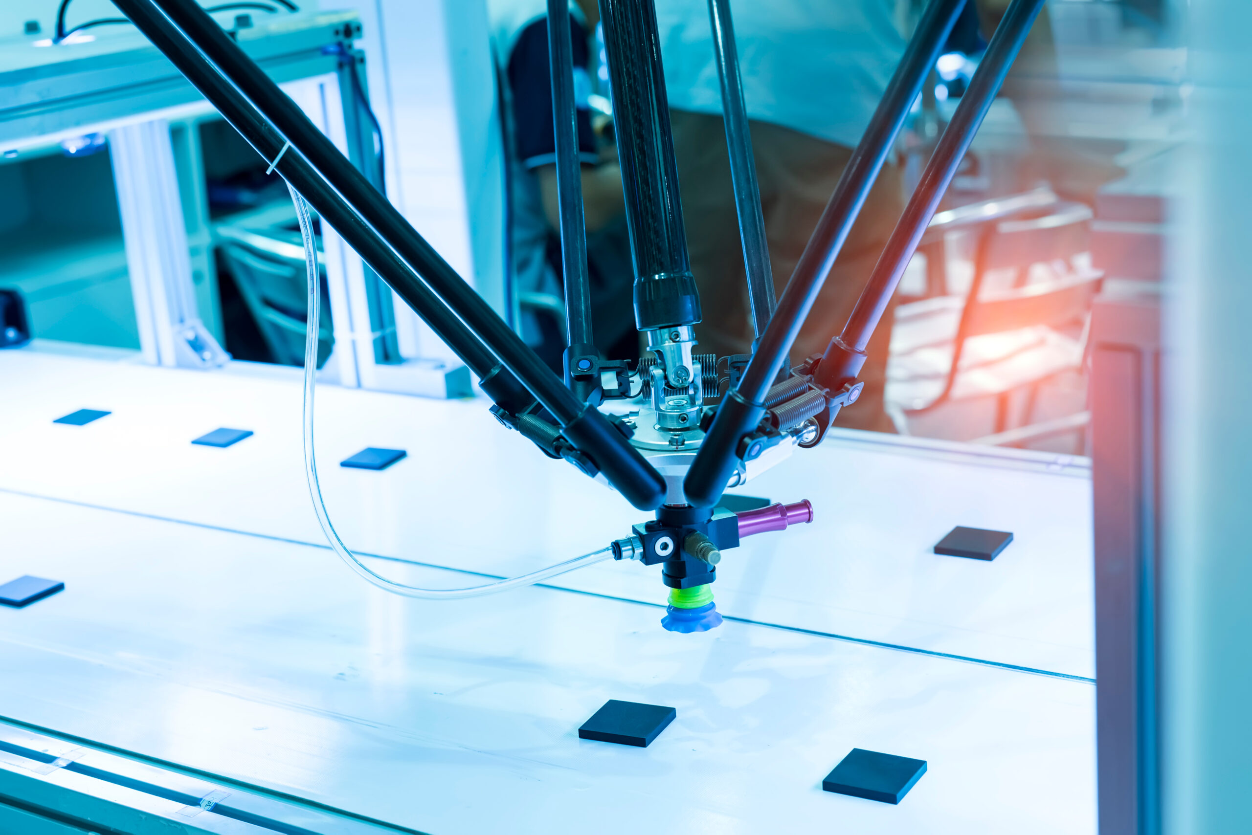 Close-up of a precision delta-style robotic arm in a medical device manufacturing clean room, positioning components on an illuminated surface. The robotic system features multiple black carbon fiber struts converging to a blue suction gripper, demonstrating advanced automation technology used in FDA-regulated medical device production environments where cybersecurity safeguards are essential throughout the manufacturing process and medical device cybersecurity.